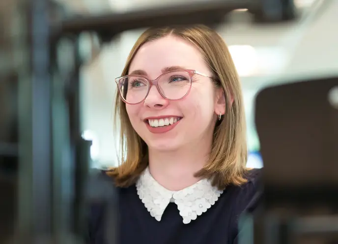 A woman wearing glasses smiling at camera whilst typing at a computer screen in an office setting.